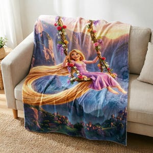 May include: A rectangular throw blanket featuring a printed illustration of a blonde character in a purple dress sitting on a floral swing. The scene includes long golden hair, a mountain landscape, and a small village at the bottom of the blanket.