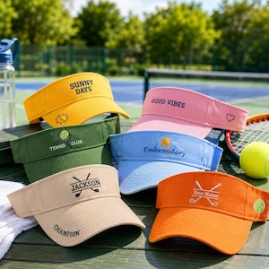 Pode incluir: Uma coleção de seis viseiras de tecido colorido com desenhos bordados. As viseiras são amarela com 'SUNNY DAYS', rosa com 'GOOD VIBES', verde com 'TENNIS CLUB', azul claro com 'Embroidery', bege com 'JACKSON' e laranja com 'YOUR NAME'.