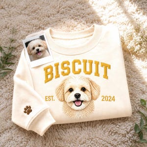 Op de afbeelding: Een crèmekleurige sweater met ronde hals, voorzien van een geborduurd portret van een lichtgekleurde hond, de tekst "BISCUIT" in het geel, "EST. 2024" in het geel en een kleine bruine pootafdruk op de mouwboord, inclusief een fotoprint.