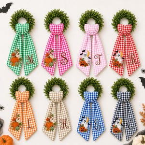 May include: Eight decorative wreaths featuring green foliage and long, hanging fabric tails in various gingham colours. Each tail displays embroidered Halloween motifs like pumpkins or ghosts and a large, stylized letter monogram.