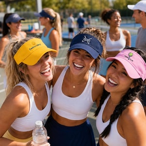 Pode incluir: Uma viseira amarela com o texto Embroidery, uma viseira azul-marinho com um gráfico de tacos de golfe cruzados e o texto Your Name, e uma viseira rosa com um gráfico de uma bola e raquete de tênis e o texto Tennis Club, usadas por pessoas.