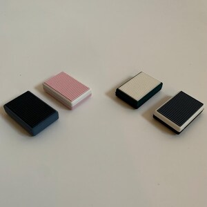 May include: Four rectangular erasers with a ribbed top surface. Two erasers feature a black top with a dark gray base, one eraser has a pink top with a white base, and one eraser has a white top with a dark gray base. Each item is a solid block.
