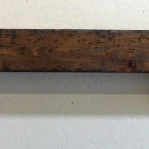 May include: A dark brown wooden shelf with two decorative brackets. The shelf is rectangular and appears to be made of reclaimed wood. The brackets are curved and add a decorative element to the shelf. The shelf is mounted on a white wall.