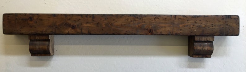 May include: A dark brown wooden shelf with two decorative brackets. The shelf is rectangular and appears to be made of reclaimed wood. The brackets are curved and add a decorative element to the shelf. The shelf is mounted on a white wall.