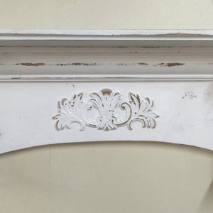 Pode incluir: Uma lareira branca com acabamento envelhecido e entalhes decorativos. A lareira apresenta uma sec&ccedil;&atilde;o central arqueada com um desenho floral e volutas decorativas nas laterais. O aspeto envelhecido confere um charme vintage &agrave; estrutura de madeira.