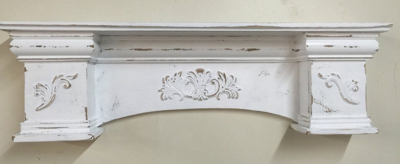 Pode incluir: Uma lareira branca com acabamento envelhecido e entalhes decorativos. A lareira apresenta uma sec&ccedil;&atilde;o central arqueada com um desenho floral e volutas decorativas nas laterais. O aspeto envelhecido confere um charme vintage &agrave; estrutura de madeira.