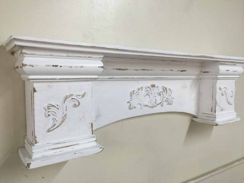 Pode incluir: Uma lareira branca com acabamento desgastado e entalhes ornamentados. A lareira apresenta um tampo retangular, molduras decorativas e desenhos florais entalhados. O acabamento desgastado revela toques de madeira, adicionando um charme r&uacute;stico.