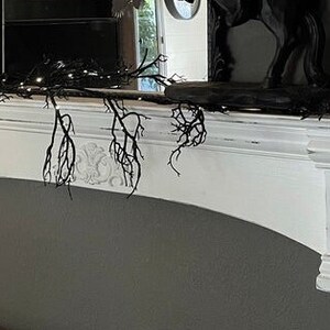 Pode incluir: Uma lareira pintada de branco com entalhes decorativos e um arco curvo. Galhos pretos e luzes de corda s&atilde;o drapeados na parte superior, criando uma est&eacute;tica g&oacute;tica. Um espelho e objetos decorativos est&atilde;o na lareira.