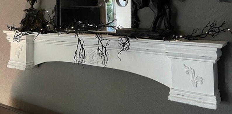Pode incluir: Uma lareira pintada de branco com entalhes decorativos e um arco curvo. Galhos pretos e luzes de corda s&atilde;o drapeados na parte superior, criando uma est&eacute;tica g&oacute;tica. Um espelho e objetos decorativos est&atilde;o na lareira.