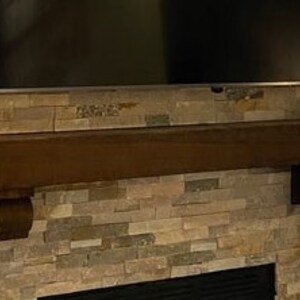 May include: A dark brown wooden mantelpiece with decorative corbels is mounted on a stone veneer wall. A large black television is mounted above the mantel. The stone veneer has a mix of brown, tan, and gray tones.