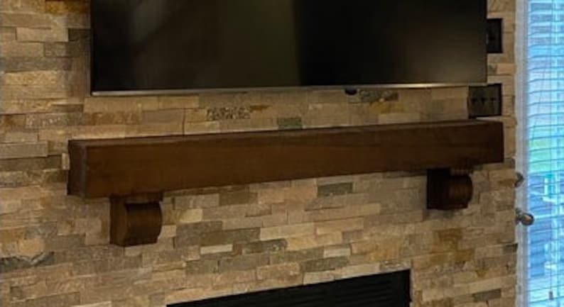 May include: A dark brown wooden mantelpiece with decorative corbels is mounted on a stone veneer wall. A large black television is mounted above the mantel. The stone veneer has a mix of brown, tan, and gray tones.