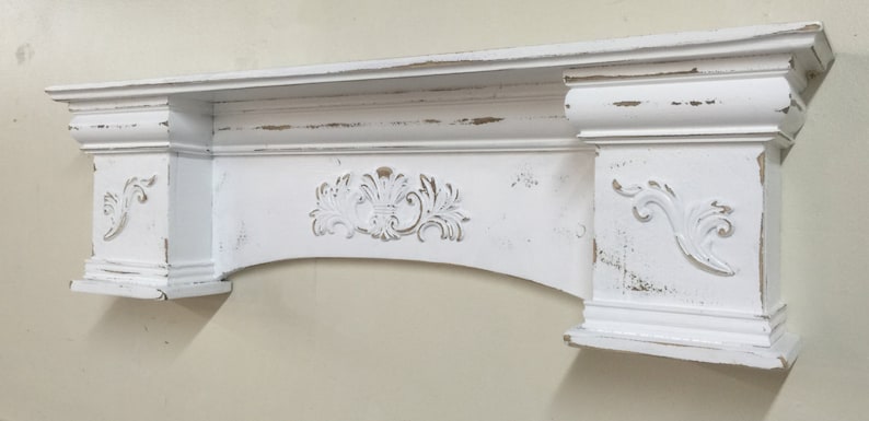 Pode incluir: Uma lareira de madeira branca desgastada com entalhes ornamentados. A lareira apresenta um design floral central e detalhes decorativos nas laterais. A pintura tem uma apar&ecirc;ncia desgastada e envelhecida, adicionando charme vintage.