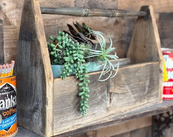 May include: A rustic wooden toolbox with a handle, filled with green succulents and other plants. The toolbox is made of weathered wood and sits on a wooden shelf. A can of coffee is visible on the left.