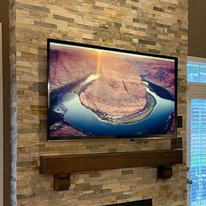 May include: A large flat-screen television mounted on a stone fireplace. The screen displays a scenic view of a river winding through a canyon. A dark wooden mantel is below the TV.