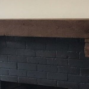 May include: A dark brown wooden fireplace mantel with decorative corbels. The mantel is mounted above a black brick fireplace. A green leafy plant is visible on the right side of the image.
