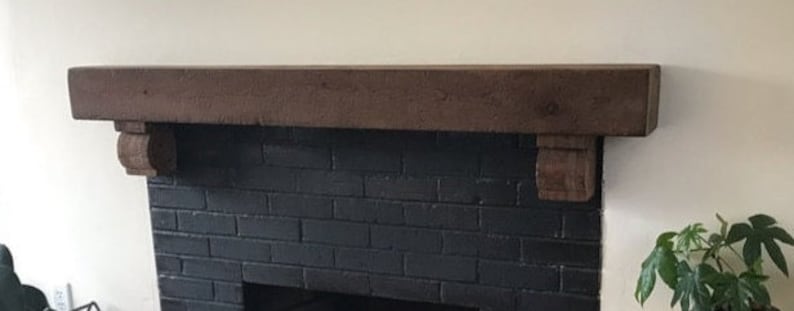 May include: A dark brown wooden fireplace mantel with decorative corbels. The mantel is mounted above a black brick fireplace. A green leafy plant is visible on the right side of the image.