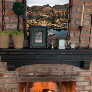May include: A black mantelpiece with decorative items against a red brick wall. The mantel holds two potted plants, a framed picture, a mirror, a clock, a small decorative item, and two candle holders with candles.
