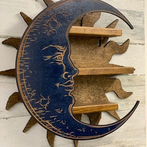 May include: A wooden wall shelf in the shape of a crescent moon with a face, painted dark blue, and a sunburst backing. The shelf has three wooden shelves for displaying small items. The sun and moon are brown.