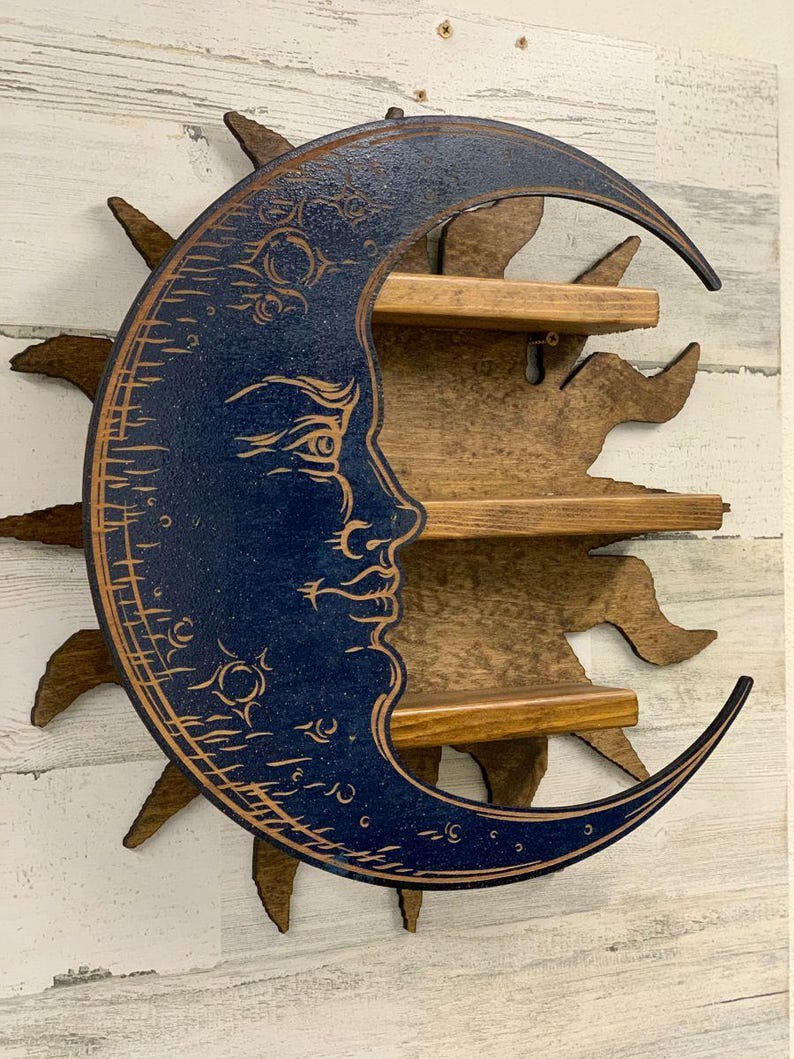 May include: A wooden wall shelf in the shape of a crescent moon with a face, painted dark blue, and a sunburst backing. The shelf has three wooden shelves for displaying small items. The sun and moon are brown.