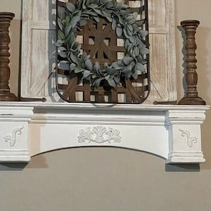 Pode incluir: Uma lareira branca com entalhes decorativos, ladeada por dois casti&ccedil;ais de madeira castanha. Uma cesta tecida com uma coroa verde pendura-se acima da lareira. Pain&eacute;is decorativos de madeira com um design geom&eacute;trico em ambos os lados.