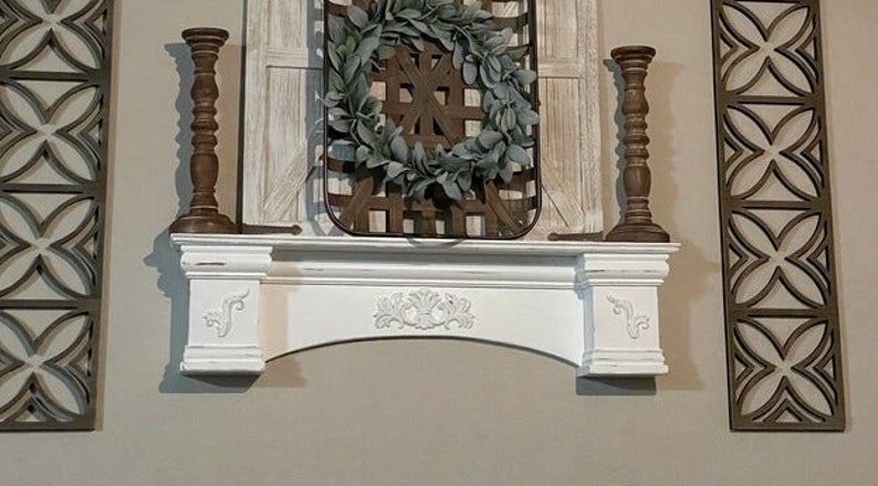 Pode incluir: Uma lareira branca com entalhes decorativos, ladeada por dois casti&ccedil;ais de madeira castanha. Uma cesta tecida com uma coroa verde pendura-se acima da lareira. Pain&eacute;is decorativos de madeira com um design geom&eacute;trico em ambos os lados.