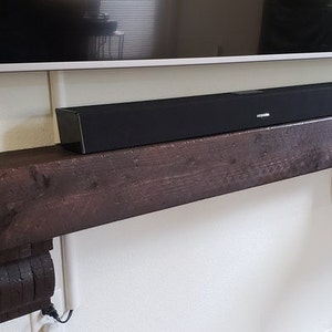May include: A black soundbar sits atop a dark brown, rustic wooden mantelpiece. The mantelpiece is supported by decorative corbels and mounted on a white wall. A television is mounted above the mantelpiece.