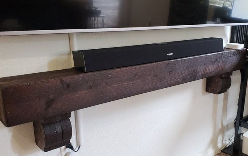 May include: A black soundbar sits atop a dark brown, rustic wooden mantelpiece. The mantelpiece is supported by decorative corbels and mounted on a white wall. A television is mounted above the mantelpiece.