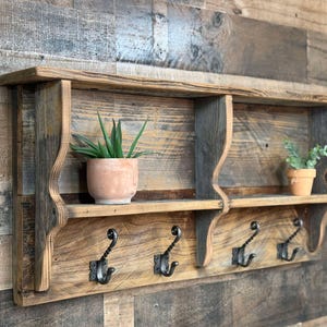 May include: Rustic wooden wall shelf with three metal hooks. The shelf has two compartments and a top shelf, with a distressed finish. Two small potted plants are displayed on the shelves. The shelf is mounted on a wooden wall.