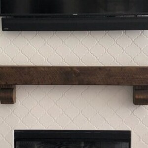 May include: A dark brown wooden mantel shelf with corbels, mounted on a white patterned wall. A black soundbar and television are mounted above the mantel. The fireplace opening is visible below.