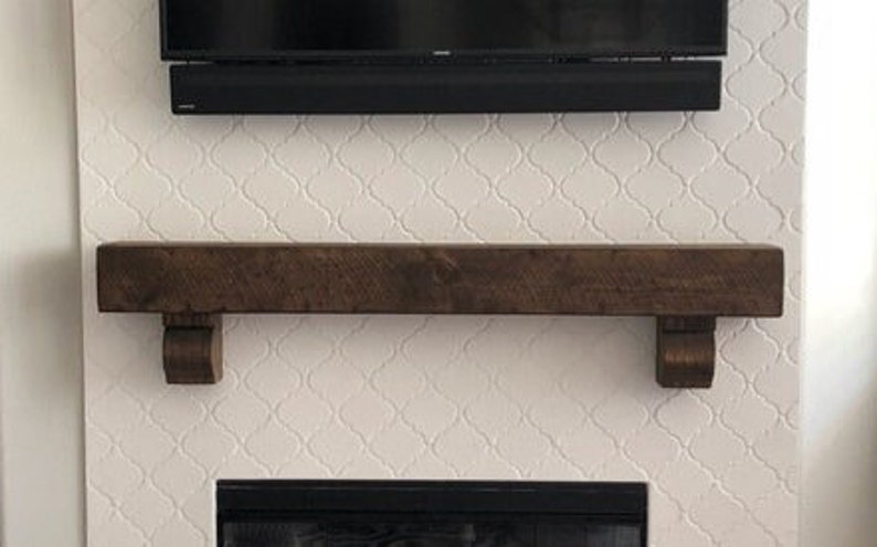 May include: A dark brown wooden mantel shelf with corbels, mounted on a white patterned wall. A black soundbar and television are mounted above the mantel. The fireplace opening is visible below.