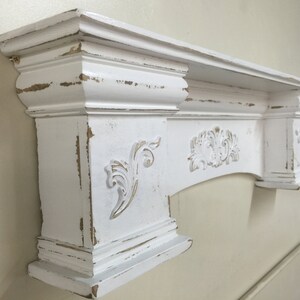 Pode incluir: Uma lareira branca com acabamento desgastado e entalhes ornamentados. A lareira apresenta um tampo retangular, molduras decorativas e detalhes florais e de rolagem. As bordas t&ecirc;m uma apar&ecirc;ncia desgastada e envelhecida.