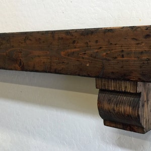 May include: A rustic, dark-stained wooden mantelpiece with corbel supports. The mantel is rectangular, with visible wood grain and a distressed finish. The corbels are carved and provide support. The overall design suggests a farmhouse or traditional aesthetic.