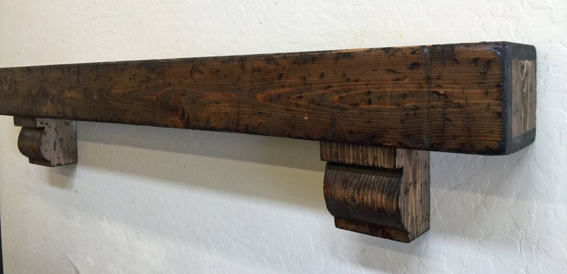 May include: A rustic, dark-stained wooden mantelpiece with corbel supports. The mantel is rectangular, with visible wood grain and a distressed finish. The corbels are carved and provide support. The overall design suggests a farmhouse or traditional aesthetic.