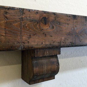 May include: A rustic, dark brown wooden mantelpiece with a distressed finish. The mantel features a rectangular beam with visible wood grain and knots, supported by a decorative corbel. The wood appears aged and weathered, adding a touch of vintage charm.