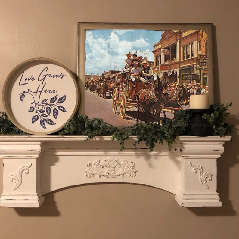 Pode incluir: Uma lareira branca com entalhes decorativos suporta uma impress&atilde;o vintage emoldurada de uma carruagem puxada por cavalos em uma rua da cidade. Uma placa redonda de madeira com o texto "Love Grows Here" e um desenho de folhas est&aacute; &agrave; esquerda. Uma vela est&aacute; em um suporte preto.