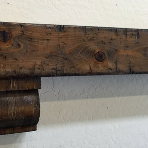 May include: A rustic, dark brown wooden mantelpiece with visible wood grain and knots. The mantel features a decorative corbel support on one end, adding a touch of elegance. The wood has a distressed finish, giving it a vintage or reclaimed appearance.
