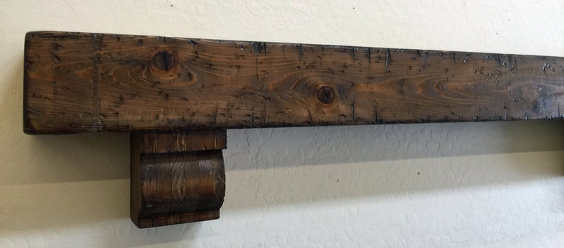 May include: A rustic, dark brown wooden mantelpiece with visible wood grain and knots. The mantel features a decorative corbel support on one end, adding a touch of elegance. The wood has a distressed finish, giving it a vintage or reclaimed appearance.