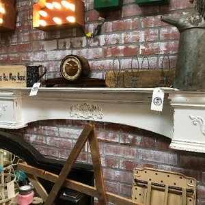 Pode incluir: Uma lareira branca desgastada com entalhes florais e um design curvo, colocada contra uma parede de tijolos vermelhos. A lareira cont&eacute;m um rel&oacute;gio vintage, uma placa de madeira que diz "We're All Mad Here!" e um jarro de metal.