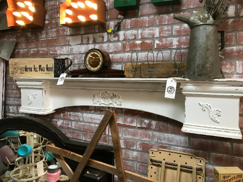 Pode incluir: Uma lareira branca desgastada com entalhes florais e um design curvo, colocada contra uma parede de tijolos vermelhos. A lareira cont&eacute;m um rel&oacute;gio vintage, uma placa de madeira que diz "We're All Mad Here!" e um jarro de metal.