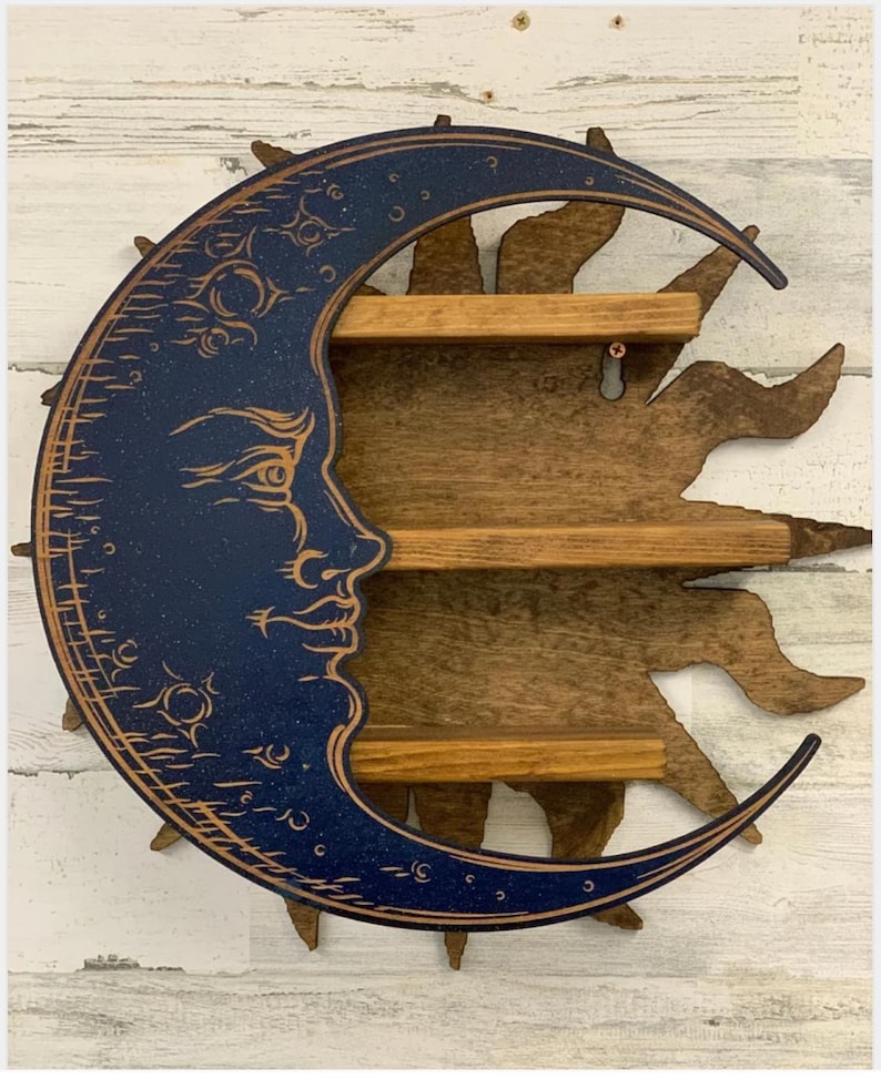 May include: A wooden wall shelf in the shape of a crescent moon with a face and a sunburst design. The moon is dark blue with gold accents, and the sun and shelves are brown. The shelf has three levels for displaying small items.