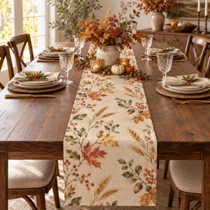 May include: A rectangular table runner made of light beige fabric featuring a printed pattern of autumn leaves, wheat stalks, red berries, and acorns. The runner is centered on a wooden dining table set with plates, glasses, and napkins.