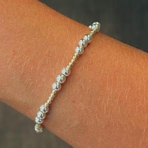 May include: A stretch bracelet featuring a repeating pattern of small, textured silver-toned metallic beads interspersed with sections of tiny, gold-toned seed beads. The design consists of alternating clusters of silver beads and gold bead segments.