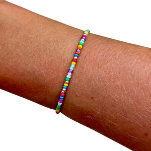 May include: A thin bracelet composed of small, round, multi-colored glass beads in shades of red, orange, yellow, green, blue, purple, and pink, interspersed with tiny metallic silver-toned spacer beads arranged in a single continuous strand.