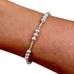 Op de afbeelding: Een elastische armband met een herhalend patroon van afwisselende metallic zilverkleurige bollen en kleinere goudkleurige kralen. Het ontwerp bestaat uit grotere, reflecterende zilveren bollen afgewisseld met groepjes van drie kleine goudkleurige kralen.