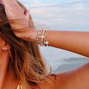 May include: A stack of five beaded bracelets worn on the wrist. The bracelets feature a combination of small metallic gold-tone beads, pink glass beads, white pearls, and a single white four-leaf clover charm made of a smooth, opaque material.