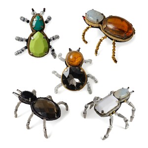 Puede incluir: Cinco broches con forma de insecto elaborados con piedras preciosas facetadas, cuentas y alambre metálico. Los diseños presentan varias combinaciones de colores, incluyendo piedras verdes, ámbar, negras y transparentes, con patas de cuentas.