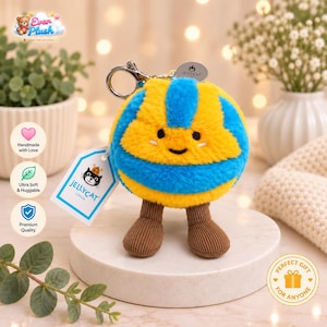 May include: A plush volleyball keychain featuring a round, fuzzy body with alternating bright yellow and blue segments. It has a smiling face, two small white marks under the eyes, and two brown corduroy legs. A Jellycat tag is attached to the silver clip.