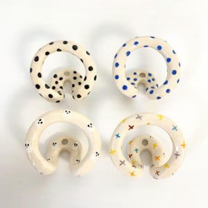 Pode incluir: Quatro ganchos de parede circulares em cerâmica com uma base de cor creme. Cada gancho apresenta um padrão único: um com pontos pretos, um com pontos azuis, um com pequenas figuras de fantasmas brancos e um com símbolos de cruz multicoloridos.