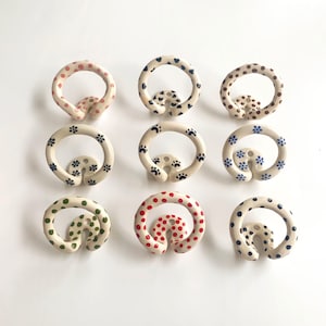 May include: Nine circular ceramic wall hooks arranged in a grid. Each hook features a unique hand-painted pattern, including various coloured polka dots, small blue flowers, and black paw prints on a cream-coloured glazed ceramic surface.