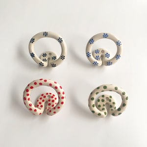 May include: Four circular ceramic wall hooks with a glossy finish. Two hooks feature small blue floral patterns on a cream base, one has red polka dots, and one has green polka dots. Each hook includes a central mounting hole for installation.